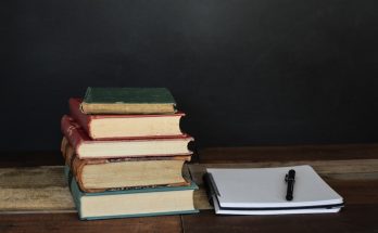 A stack of vintage books beside a blank notebook and a pen on a wooden surface