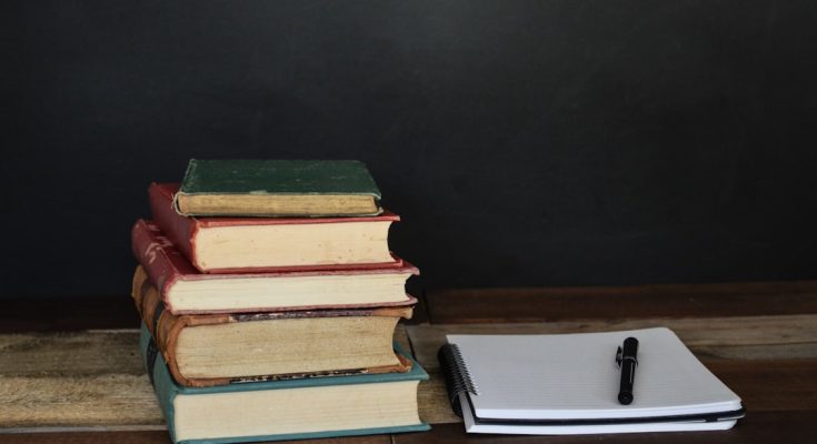 A stack of vintage books beside a blank notebook and a pen on a wooden surface